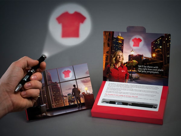Hand holding a small flashlight that projects a red shirt icon onto a wall, next to a branded direct mail package featuring a city skyline image, a woman in a red shirt, and a pen, highlighting Independent Health’s RedShirt® Treatment campaign materials.